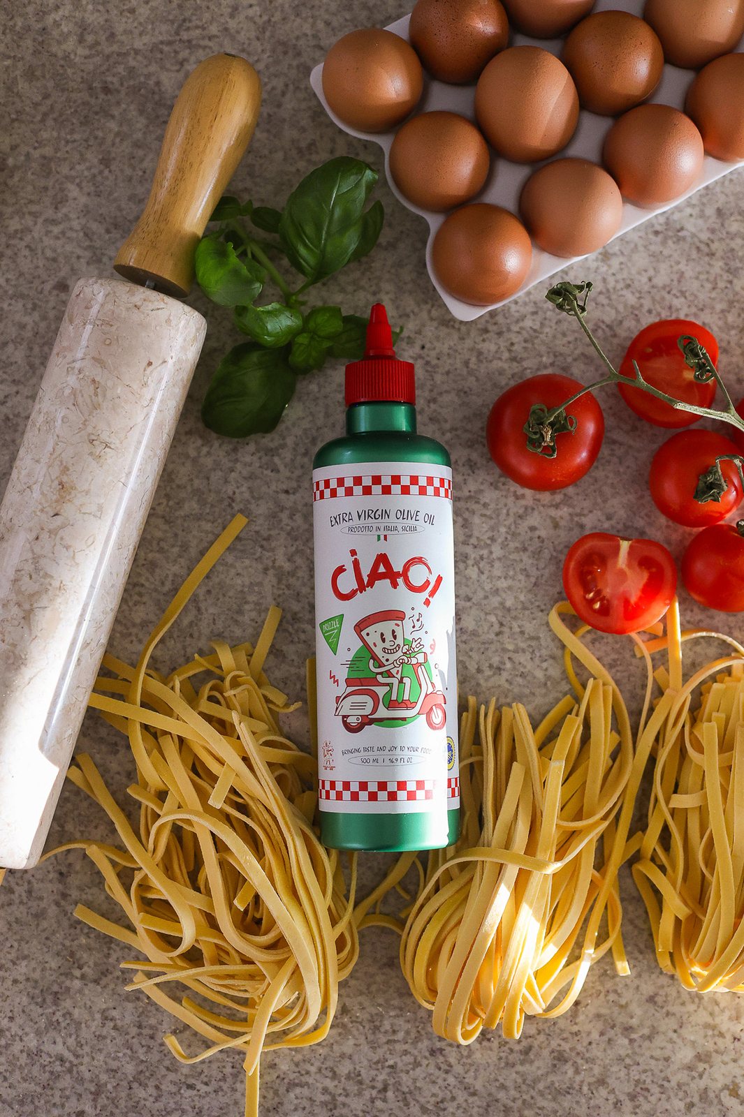 CIAO! Extra Virgin Olive Oil bottle with fresh pasta, basil, eggs and tomatoes on a kitchen table