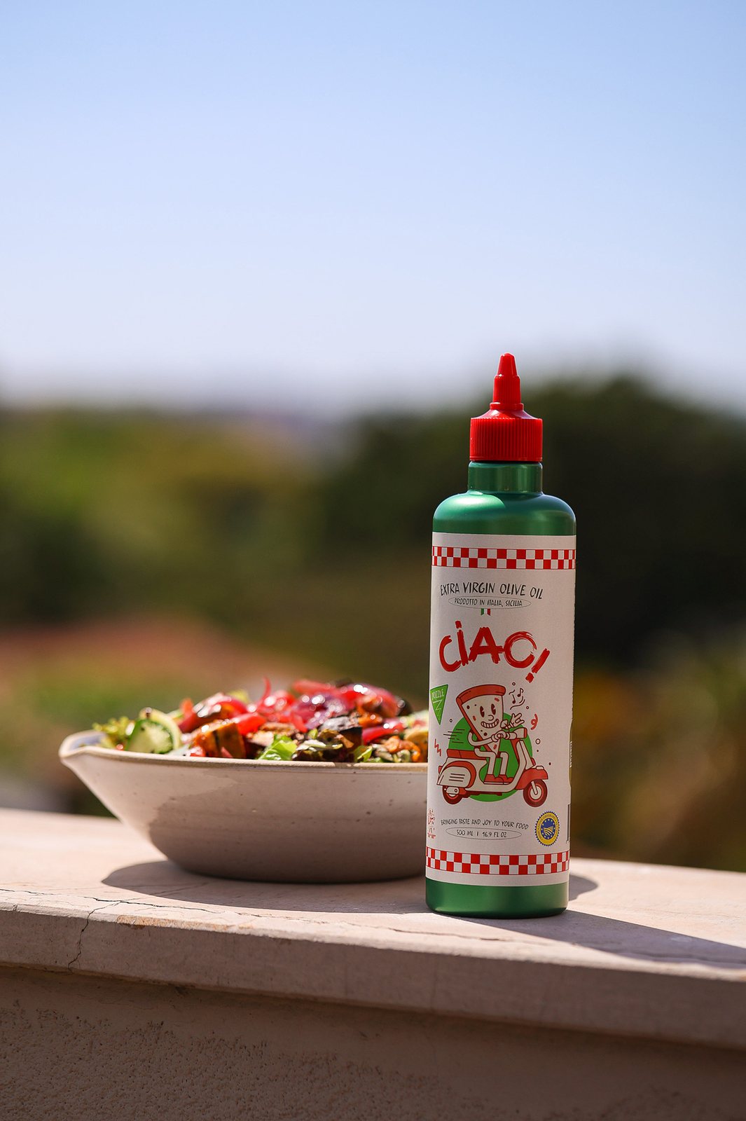 CIAO! Extra Virgin Olive Oil bottle beside a fresh Sicilian salad with a sunlit countryside backdrop